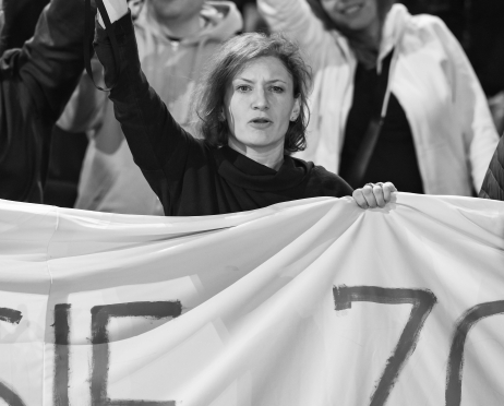 Czarno-białe zdjęcie. Martyna Krzysztofik stoi wśród protestujących w teatrze. Jedną ręką unosi megafon, w drugiej trzyma duży baner z odręcznym napisem. W tle widać inne osoby z uniesionymi rękami, wspierające protest.
